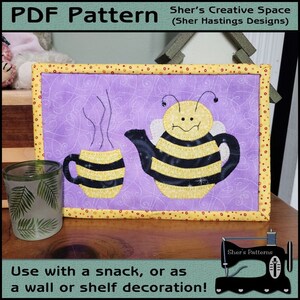 May include: A purple and yellow quilted wall hanging featuring a bee-shaped teapot and a cup with steam coming from it. The text "Sher's Creative Space (Sher Hastings Designs)" is at the top of the image. The text "Use with a snack, or as a wall or shelf decoration!" is at the bottom of the image.