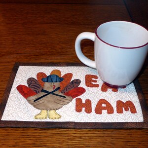 Peut inclure: Un dessous de tasse matelass&eacute; brun et blanc avec un motif de dinde et les mots "Eat Ham". Une tasse blanche avec un bord marron est pos&eacute;e sur le dessous de tasse.