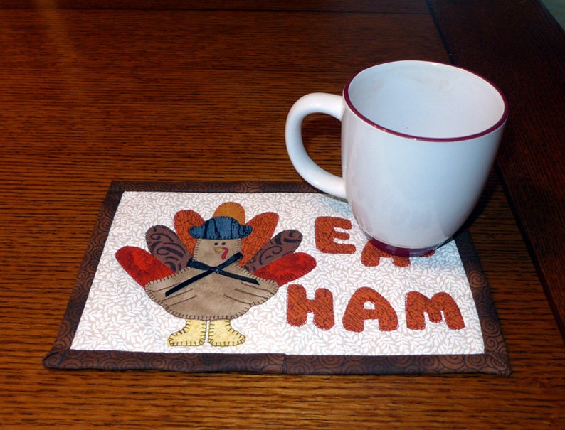 Peut inclure: Un dessous de tasse matelass&eacute; brun et blanc avec un motif de dinde et les mots "Eat Ham". Une tasse blanche avec un bord marron est pos&eacute;e sur le dessous de tasse.