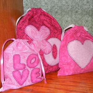 May include: Three pink fabric drawstring bags with different heart designs. The bags are decorated with pink and white swirls and the word "LOVE" is stitched on one bag.