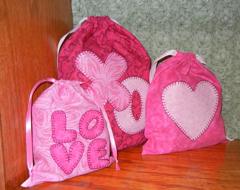 May include: Three pink fabric drawstring bags with different heart designs. The bags are decorated with pink and white swirls and the word "LOVE" is stitched on one bag.