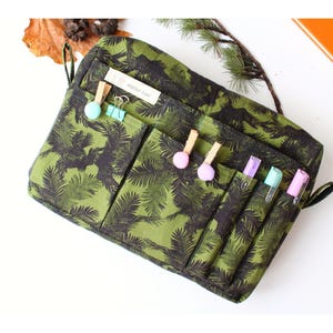 Può includere: Organizer verde e nero a fantasia con più tasche. L'organizer ha un design a foglie di palma e include piccole clip e penne. Il testo "Atelier Lini" è visibile su una piccola etichetta. Progettato per cancelleria e forniture per ufficio.