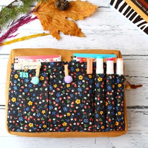 May include: A brown canvas and floral print organizer with multiple pockets. The organizer has a black background with a colorful floral pattern. It holds notebooks, pens, and markers. The organizer is designed for stationery and art supplies.