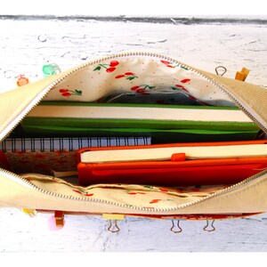 May include: Beige canvas pencil case with a cherry print lining, open to reveal notebooks and a red journal. The case is zippered and sits on a white wooden surface. Several binder clips are attached to the case.