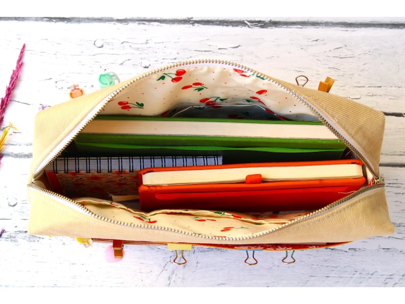 May include: Beige canvas pencil case with a cherry print lining, open to reveal notebooks and a red journal. The case is zippered and sits on a white wooden surface. Several binder clips are attached to the case.