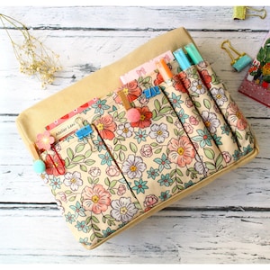 May include: A rectangular fabric desk organizer featuring a cream background with a repeating floral pattern in shades of pink, teal, and green. The organizer has multiple vertical pockets containing various pens, clips, and small paper items.