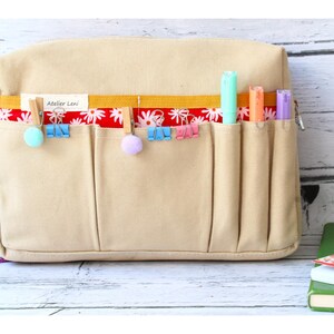 May include: Beige canvas organizer with multiple pockets, accented with a red floral patterned ribbon and a yellow band. Includes colorful markers and clips. The text "Atelier Leni" is visible on the ribbon. Ideal for stationery and craft supplies.