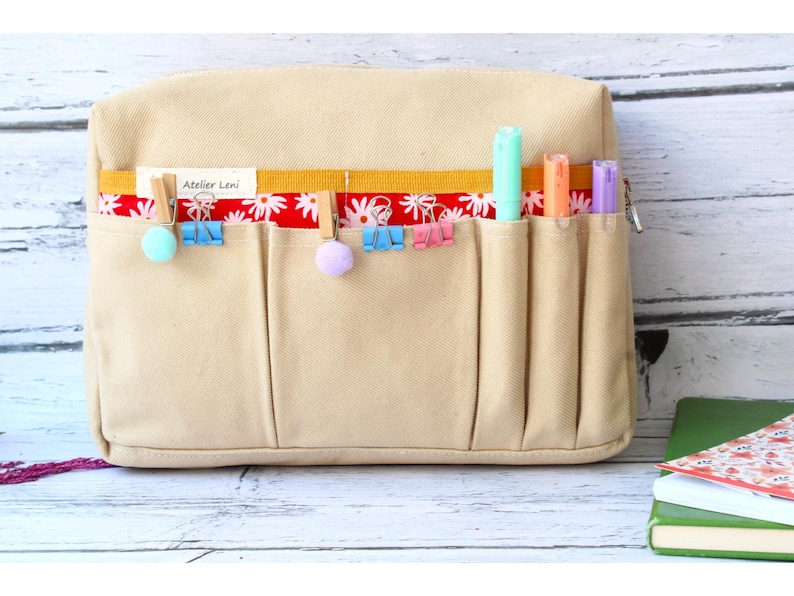 May include: Beige canvas organizer with multiple pockets, accented with a red floral patterned ribbon and a yellow band. Includes colorful markers and clips. The text "Atelier Leni" is visible on the ribbon. Ideal for stationery and craft supplies.