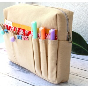 May include: Beige canvas pencil case with a silver zipper and multiple pockets. The pockets hold colorful pens and decorative clips. A red and white floral pattern accents the top of the pockets.