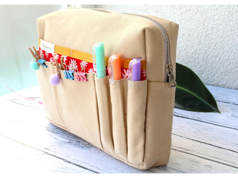 May include: Beige canvas pencil case with a silver zipper and multiple pockets. The pockets hold colorful pens and decorative clips. A red and white floral pattern accents the top of the pockets.