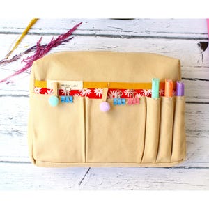 May include: Beige canvas pencil case with multiple pockets. A red and white floral ribbon and yellow ribbon are attached to the top. The case has colorful binder clips, pom-poms, and highlighters. The text "Atelier Leni" is visible on a small tag.