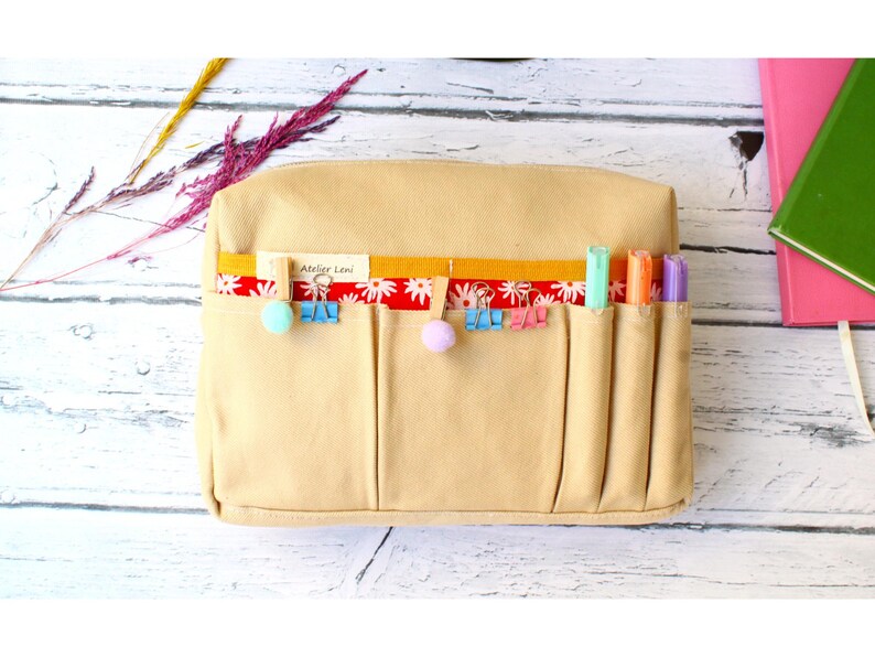 May include: Beige canvas pencil case with multiple pockets. A red and white floral ribbon and yellow ribbon are attached to the top. The case has colorful binder clips, pom-poms, and highlighters. The text "Atelier Leni" is visible on a small tag.
