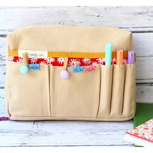 May include: Beige canvas organizer with multiple pockets, featuring a red floral patterned accent. The organizer has colorful clips and pens in the pockets. The text "Atelier Leni" is visible on the front.