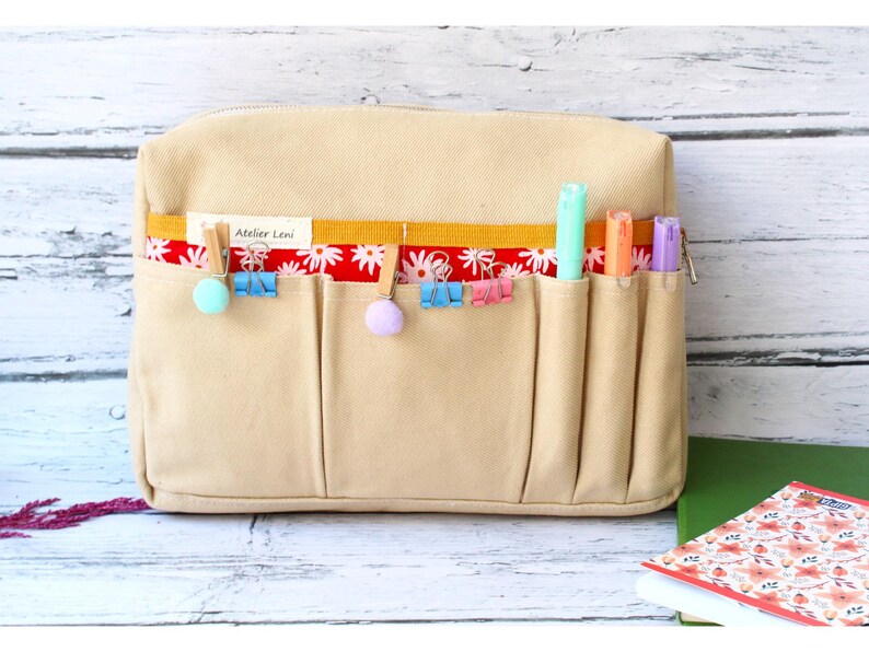 May include: Beige canvas organizer with multiple pockets, featuring a red floral patterned accent. The organizer has colorful clips and pens in the pockets. The text "Atelier Leni" is visible on the front.