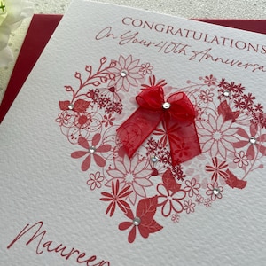 May include: A white anniversary card with a red floral heart design and a red ribbon bow. The card reads "Congratulations On Your 40th Anniversary" in red script. The name "Maureen" is also written on the card.