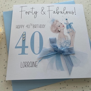 May include: A light blue birthday card with the text "Forty & Fabulous!" and "Happy 40th Birthday". The card features an illustration of a woman holding a champagne bottle and a large blue bow.