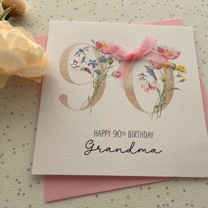 May include: A white greeting card for a 90th birthday with the word "Grandma" written in cursive. The number 90 is decorated with floral illustrations and a pink ribbon. The card is on top of a pink envelope.