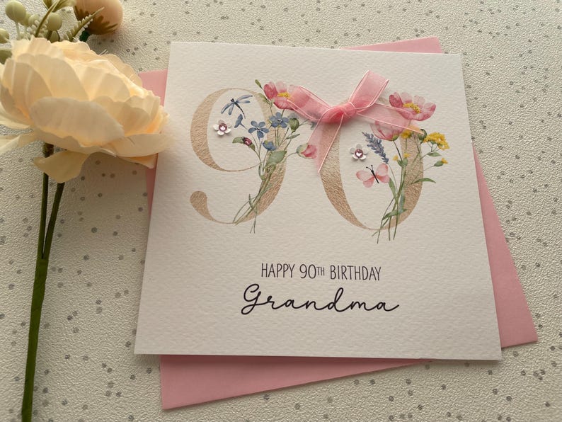 May include: A white greeting card for a 90th birthday with the word "Grandma" written in cursive. The number 90 is decorated with floral illustrations and a pink ribbon. The card is on top of a pink envelope.