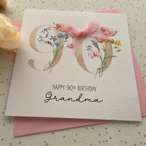 May include: A white birthday card for a 90th birthday with the words "Happy 90th Birthday Grandma" in black script. The number 90 is decorated with flowers and a pink bow. The card is on a pink envelope.