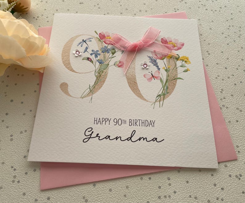 May include: A white birthday card for a 90th birthday with the words "Happy 90th Birthday Grandma" in black script. The number 90 is decorated with flowers and a pink bow. The card is on a pink envelope.