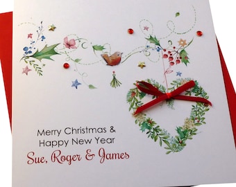 May include: A white Christmas card with a red envelope. The card features a watercolor illustration of a heart-shaped wreath with a red ribbon, a robin, and holly. The text on the card reads "Merry Christmas & Happy New Year Sue, Roger & James".