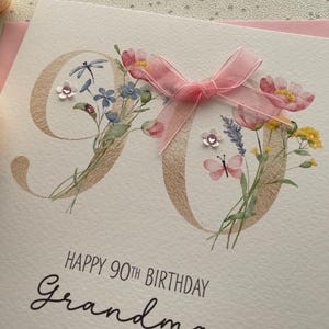 May include: A birthday card with the number 90 in gold, decorated with watercolor flowers and a pink ribbon. The card reads "Happy 90th Birthday Grandma" in elegant script.