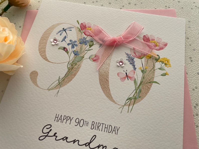 May include: A birthday card with the number 90 in gold, decorated with watercolor flowers and a pink ribbon. The card reads "Happy 90th Birthday Grandma" in elegant script.