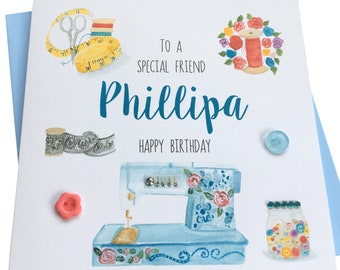May include: A birthday card with a sewing theme. The card features a blue sewing machine with floral accents, a spool of thread, a measuring tape, scissors, a jar of buttons, and a ribbon. The card is addressed to "Phillipa" and says "To a special friend, Happy Birthday".