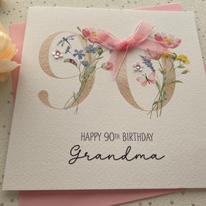May include: A white birthday card with the number 90 in gold, adorned with watercolor flowers and a pink ribbon. The card reads "Happy 90th Birthday Grandma" in elegant script, set against a pink envelope.