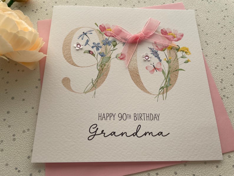 May include: A white birthday card with the number 90 in gold, adorned with watercolor flowers and a pink ribbon. The card reads "Happy 90th Birthday Grandma" in elegant script, set against a pink envelope.