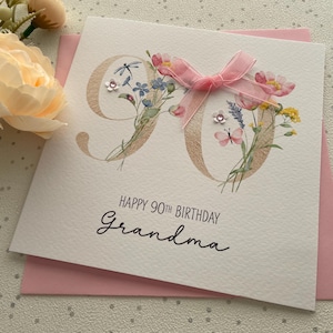 May include: A white card for a 90th birthday, with the words 'Happy 90th Birthday Grandma'. The number 90 is decorated with flowers and a pink ribbon. The card is on a pink envelope.