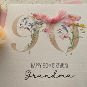 May include: A handmade birthday card for a 90th birthday. The card features a gold-colored "90" with floral accents, a pink ribbon, and the words "Happy 90th Birthday Grandma" in black script.
