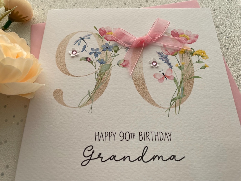 May include: A handmade birthday card for a 90th birthday. The card features a gold-colored "90" with floral accents, a pink ribbon, and the words "Happy 90th Birthday Grandma" in black script.