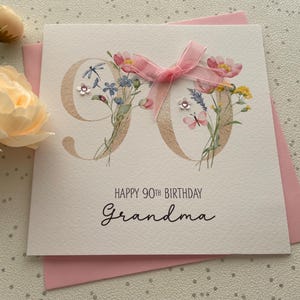 May include: A square greeting card with a pink envelope. The card features the number "90" in a light brown color, decorated with watercolor flowers and a pink ribbon. The text "HAPPY 90TH BIRTHDAY Grandma" is printed below.