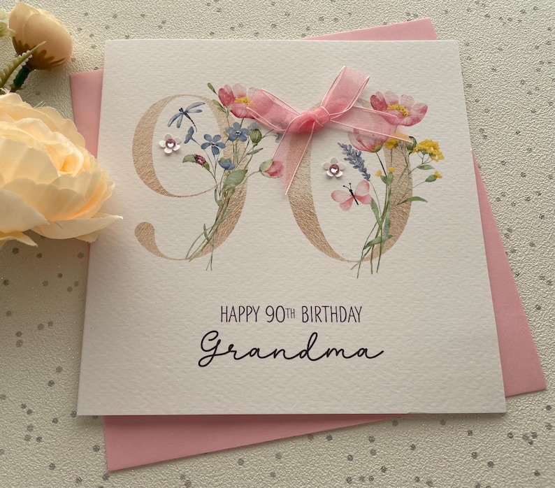 May include: A square greeting card with a pink envelope. The card features the number "90" in a light brown color, decorated with watercolor flowers and a pink ribbon. The text "HAPPY 90TH BIRTHDAY Grandma" is printed below.
