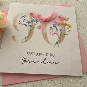 May include: A white greeting card with the number 90 in gold, decorated with watercolor flowers and a pink ribbon. The card reads "HAPPY 90TH BIRTHDAY Grandma" in black script. A pink envelope is visible behind the card.