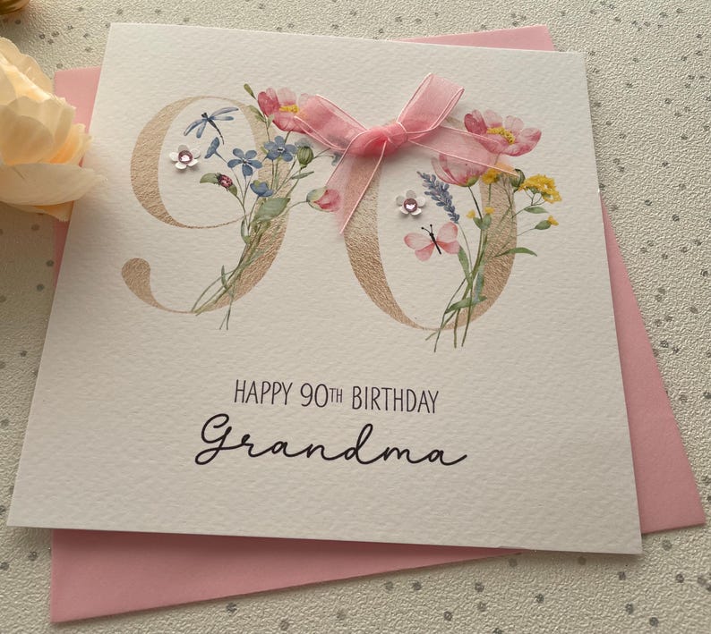 May include: A white greeting card with the number 90 in gold, decorated with watercolor flowers and a pink ribbon. The card reads "HAPPY 90TH BIRTHDAY Grandma" in black script. A pink envelope is visible behind the card.