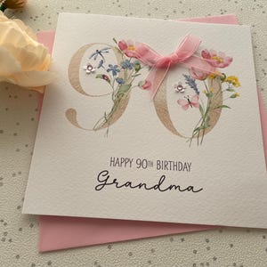 May include: A white birthday card with the number 90 in gold, decorated with flowers and a pink bow. The card reads "Happy 90th Birthday Grandma" in black script, with a pink envelope underneath.
