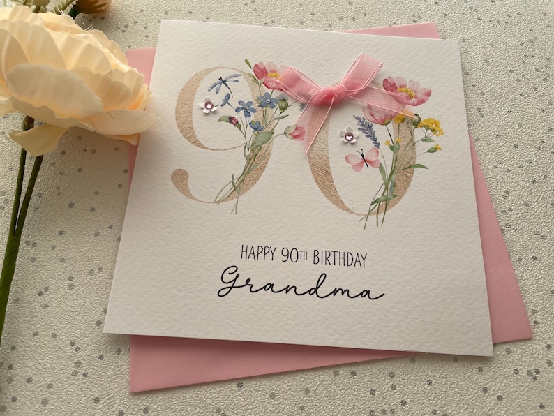 May include: A white birthday card with the number 90 in gold, decorated with flowers and a pink bow. The card reads "Happy 90th Birthday Grandma" in black script, with a pink envelope underneath.