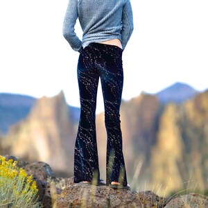 Puede incluir: Una mujer con un su&eacute;ter gris y pantalones acampanados azul oscuro con un estampado floral blanco. Lleva un sombrero marr&oacute;n con un adorno de plumas y est&aacute; de pie sobre una superficie rocosa.