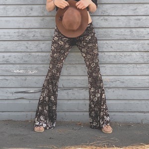 Op de afbeelding: Een vrouw draagt een zwart topje met een off-the-shoulder-look en een bruine en witte bloemenprint bellbottom broek. Ze houdt een bruine fedora-hoed voor zich.