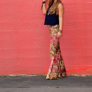 May include: A woman wearing a dark blue sleeveless top and patterned pants stands in front of a red wall. She is wearing a necklace and bracelets.
