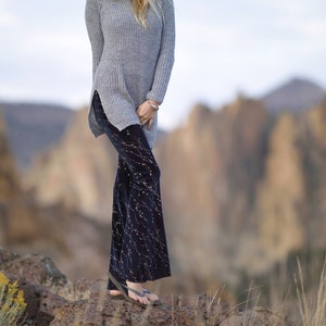 Puede incluir: Una mujer con un su&eacute;ter gris, un sombrero fedora marr&oacute;n y pantalones con estampado negro y dorado se para sobre una superficie rocosa. El fondo est&aacute; desenfocado y muestra un paisaje rocoso.