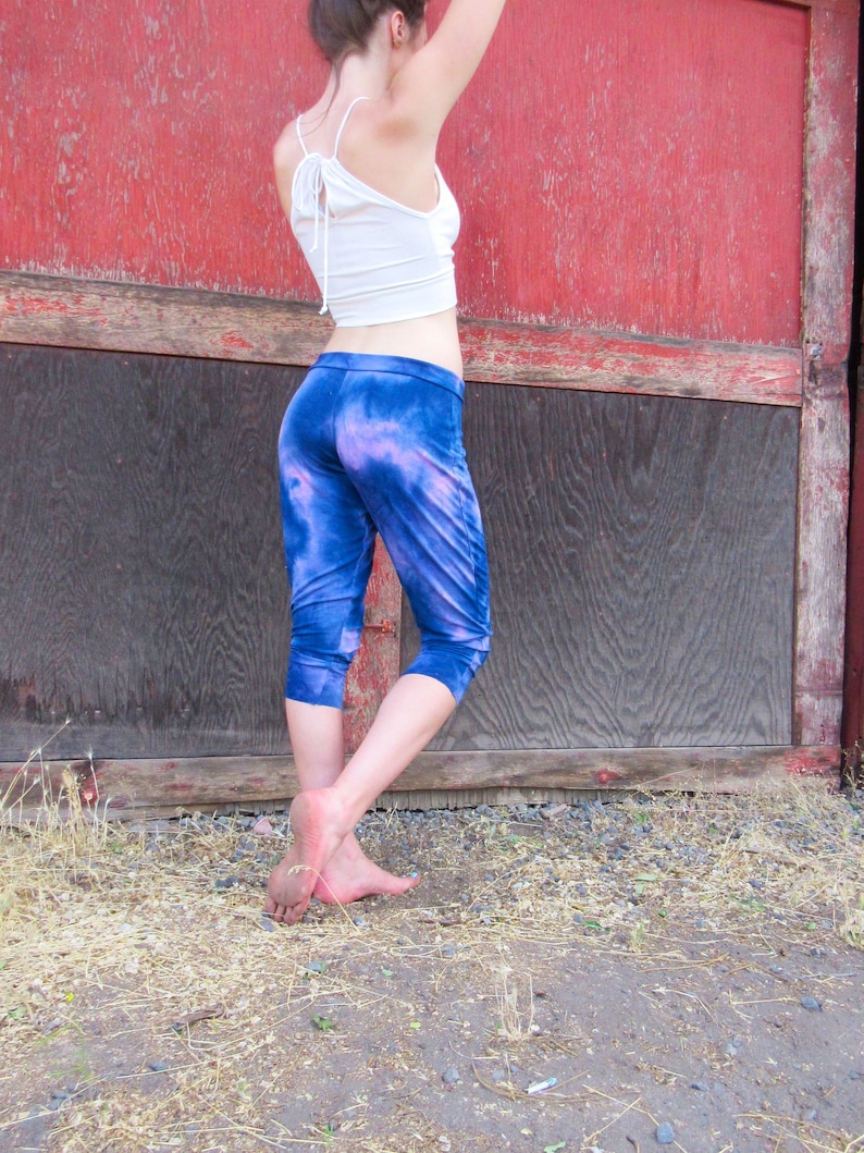 May include: A person wearing a white crop top and blue tie-dye pants. The person is standing in front of a red and black wooden door.