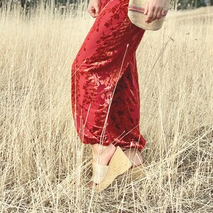 May include: A woman wearing red velvet pants with a floral pattern and a woven bag. She is standing in a field of tall, dry grass.