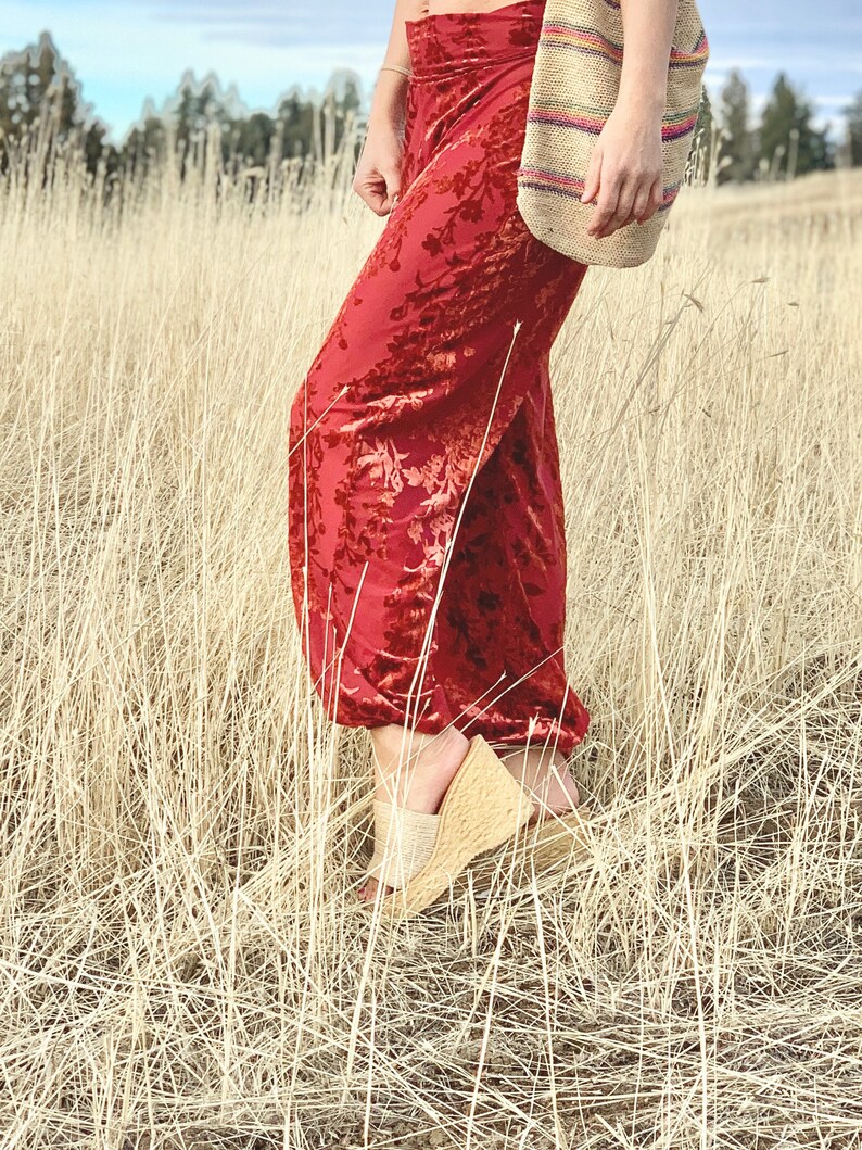 May include: A woman wearing red velvet pants with a floral pattern and a woven bag. She is standing in a field of tall, dry grass.