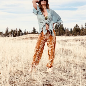 May include: A woman wearing a light blue kimono with a green bra peeking out, brown wide-brimmed hat, and floral print pants. She is standing in a field of tall, dry grass.
