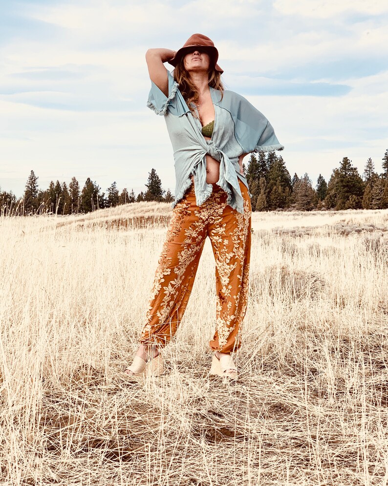 May include: A woman wearing a light blue kimono with a green bra peeking out, brown wide-brimmed hat, and floral print pants. She is standing in a field of tall, dry grass.