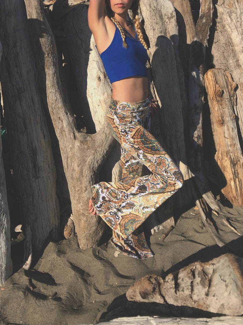 May include: A woman wearing a blue crop top and patterned bell bottom pants stands in front of a wall of driftwood.