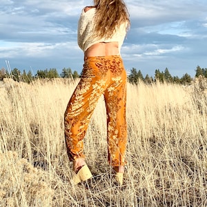 May include: A woman wearing a white sweater, brown hat, and orange floral print pants stands in a field of tall grass.